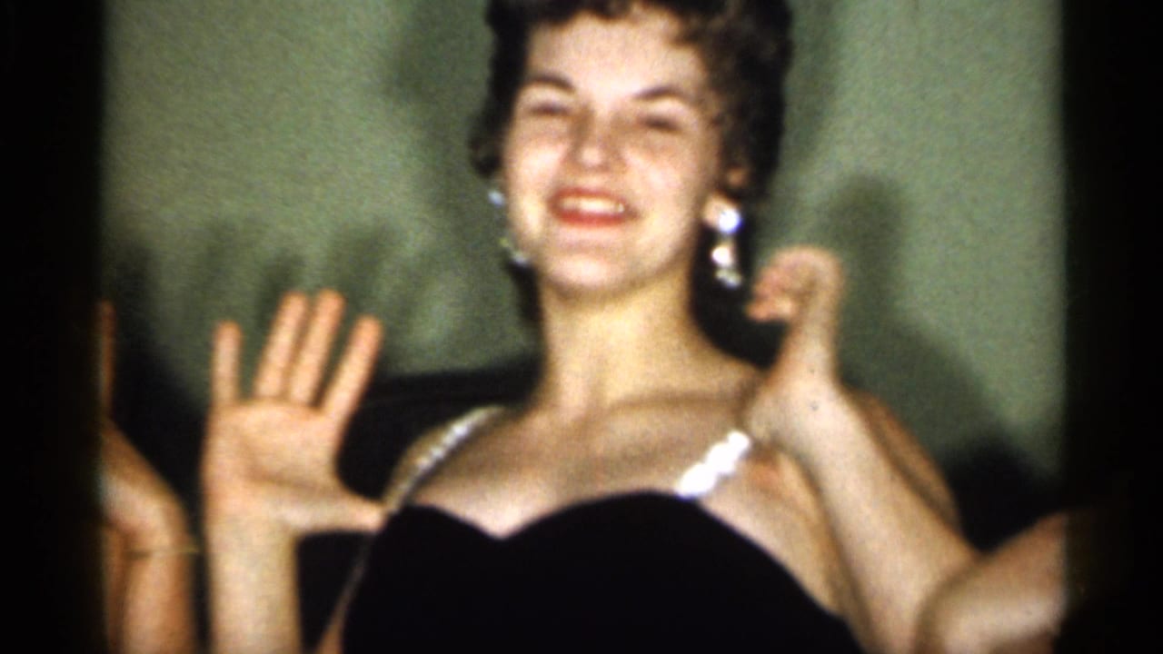 A young woman in a dark dress with white trim smiles with a raised hand in 1958 New Jersey home-movie footage.