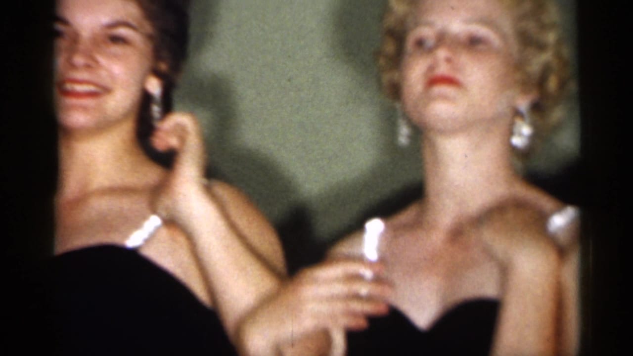 Two young women in dark dresses with white trim stand close together indoors in 1958 New Jersey home-movie footage.