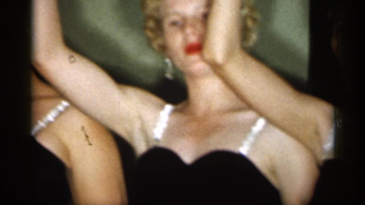 1958 New Jersey home-movie frame showing two adult women in black dresses with white straps, red lipstick, earrings, and raised arms.