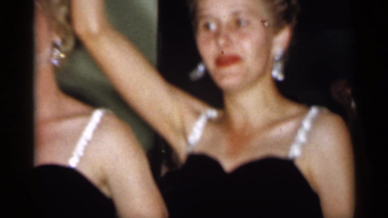 A woman in a black dress faces the camera while arms are raised around her.