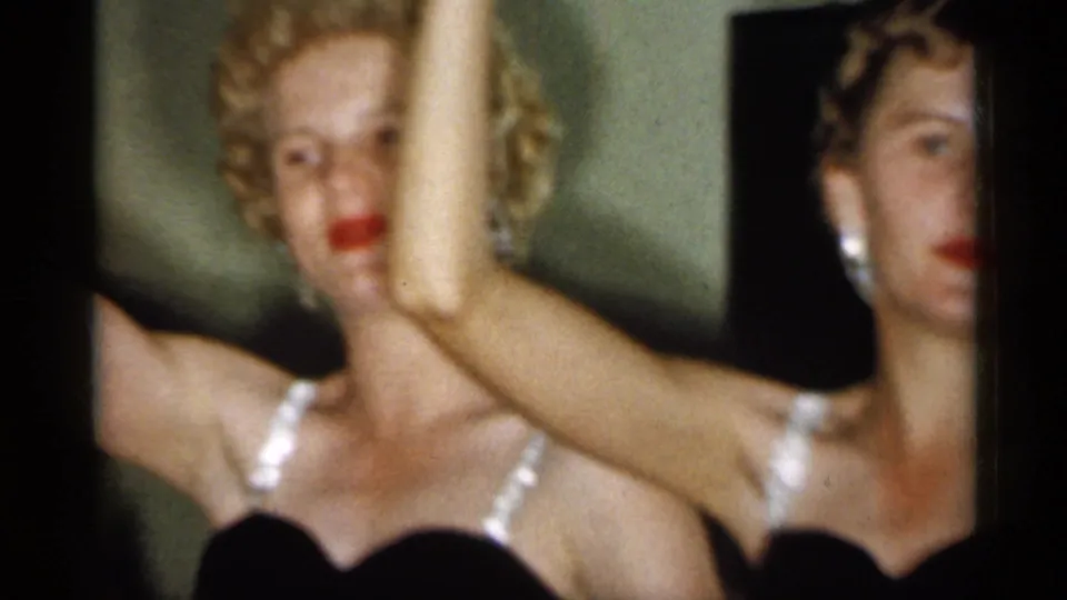 1958 New Jersey home-movie frame of two adult women in dark formal dresses, one centered and one partly hidden, both within a raised-arm pose.