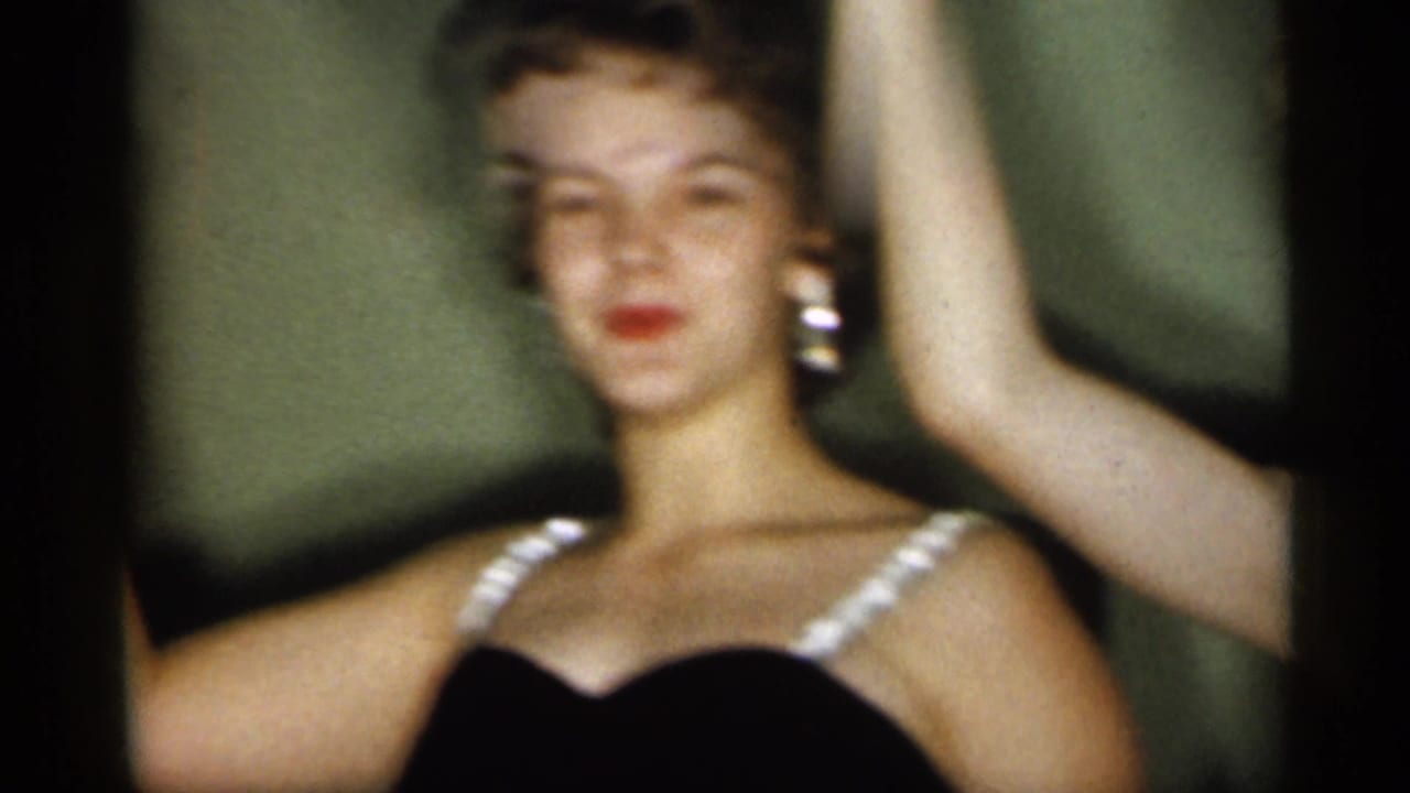 1958 New Jersey frame showing an adult woman in a black dress with white straps and earrings, with another person's raised arm at the edge.