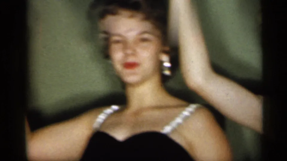 1958 New Jersey frame showing an adult woman in a black dress with white straps and earrings, with another person's raised arm at the edge.