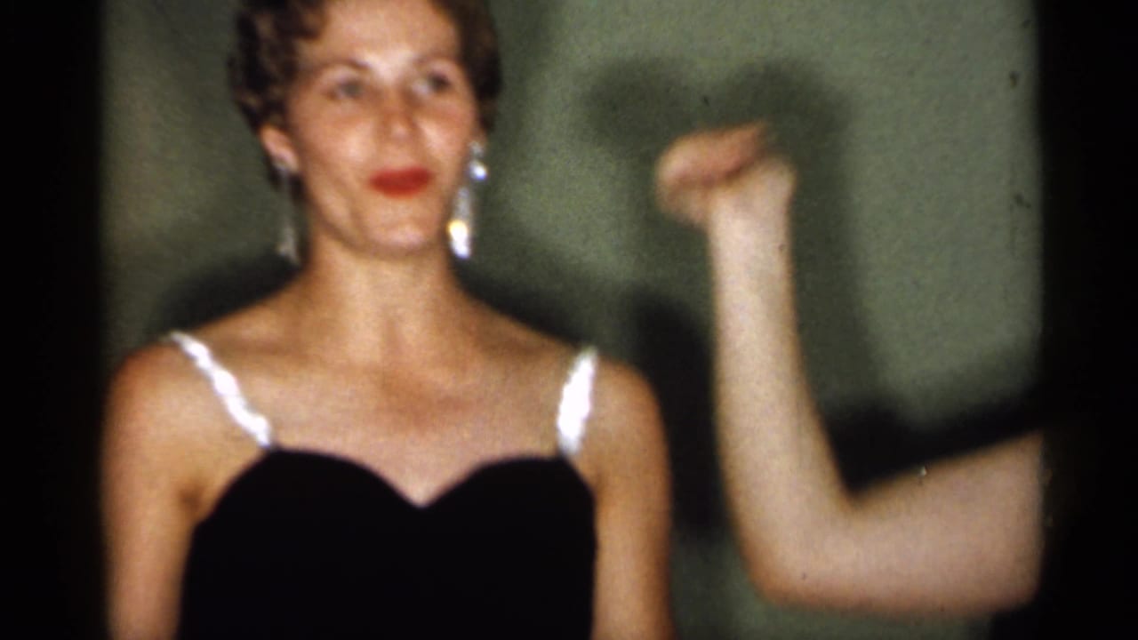 In supported 1958 New Jersey home-movie footage, a young woman in a black evening dress with white straps raises her arm indoors.