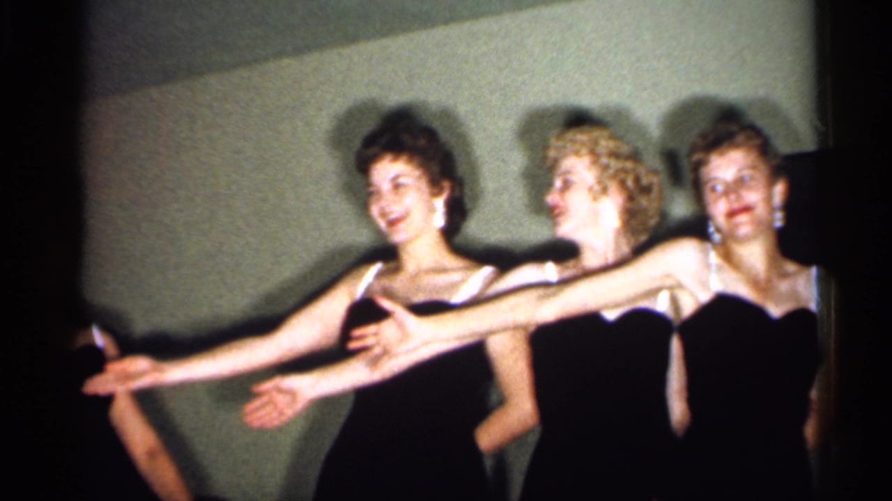 Women in formal black dresses extend their arms in a synchronized movement during the indoor 1958 New Jersey routine.