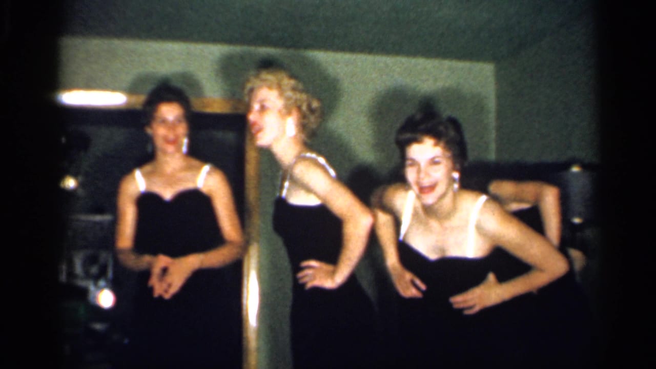 Three young women in black dresses pose in a dim room while another figure is partly visible behind them.