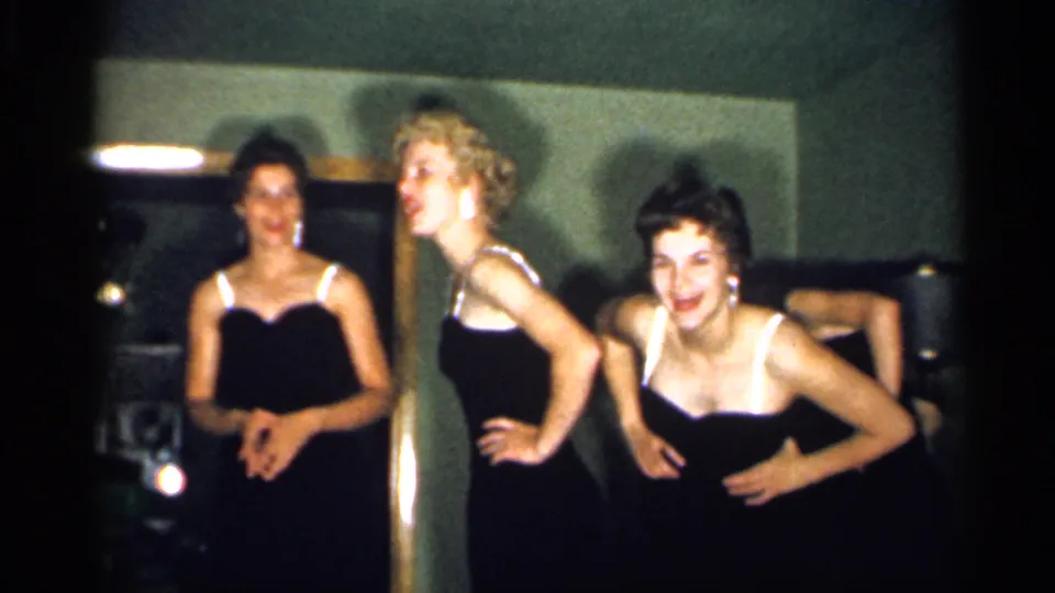 In a 1958 New Jersey home-movie frame, young women in black dresses with white straps pose and smile in a dim private interior.