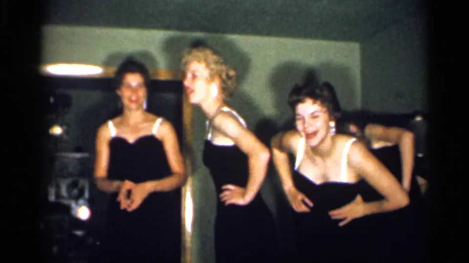 Young women in black dresses with white straps pose playfully toward the camera in archival 1958 New Jersey home-movie footage.