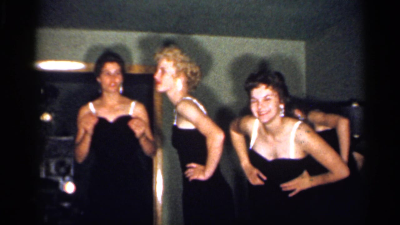 A 1958 New Jersey home-movie frame shows young women in coordinated dark dresses gesturing and posing in a private interior.