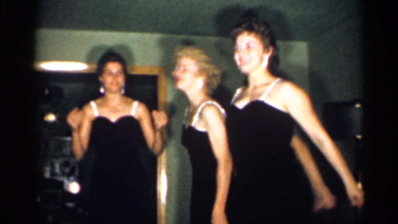 Four young women in black dresses appear to dance or sway together in a dim New Jersey interior captured on 8mm film in 1958.