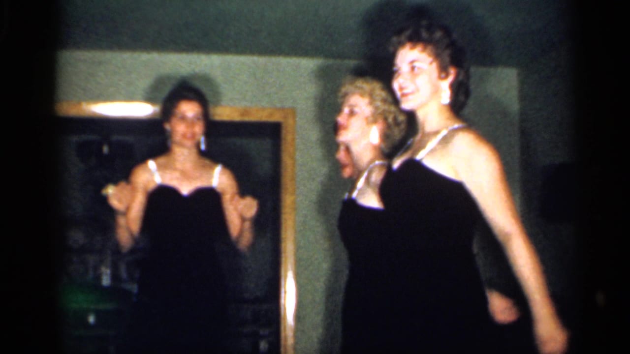Young women in black evening dresses perform in a dim living room.