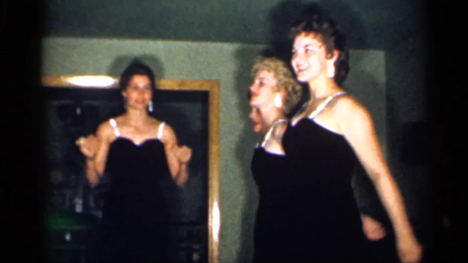 In a 1958 New Jersey home interior, young women in black evening dresses gesture during an informal performance.