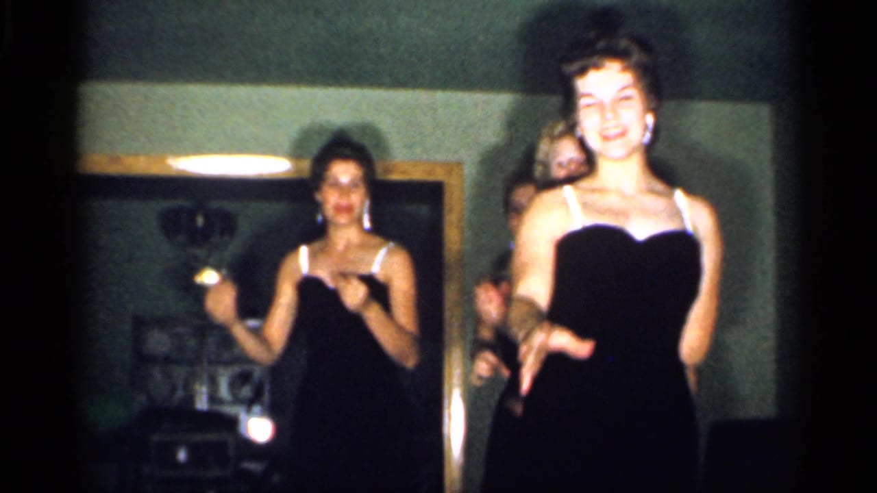 A smiling performer in a black dress moves near the camera as other women continue the routine in the living room.