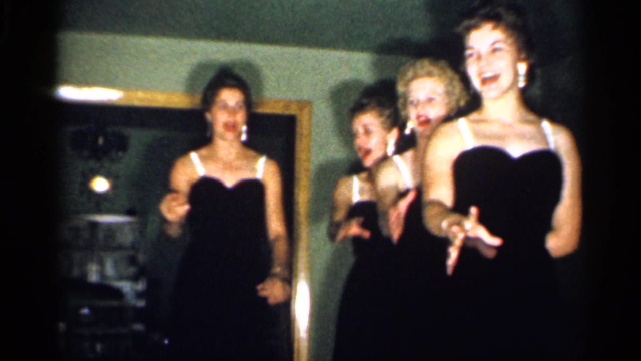The performers cluster together in the 1958 home-movie frame, smiling and gesturing in matching dark dresses.