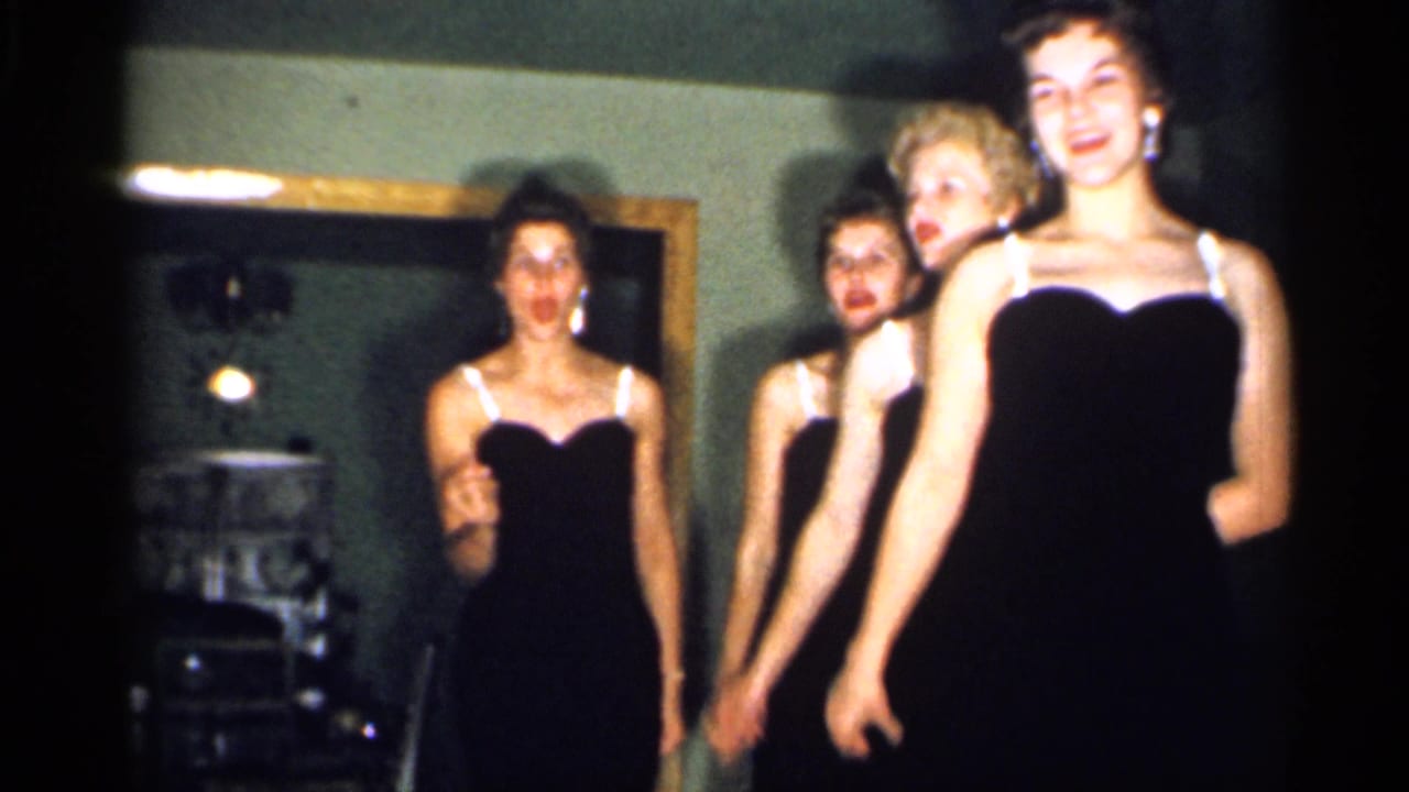 Four young women stand close together in black evening dresses during a New Jersey home performance documented on 8mm film.