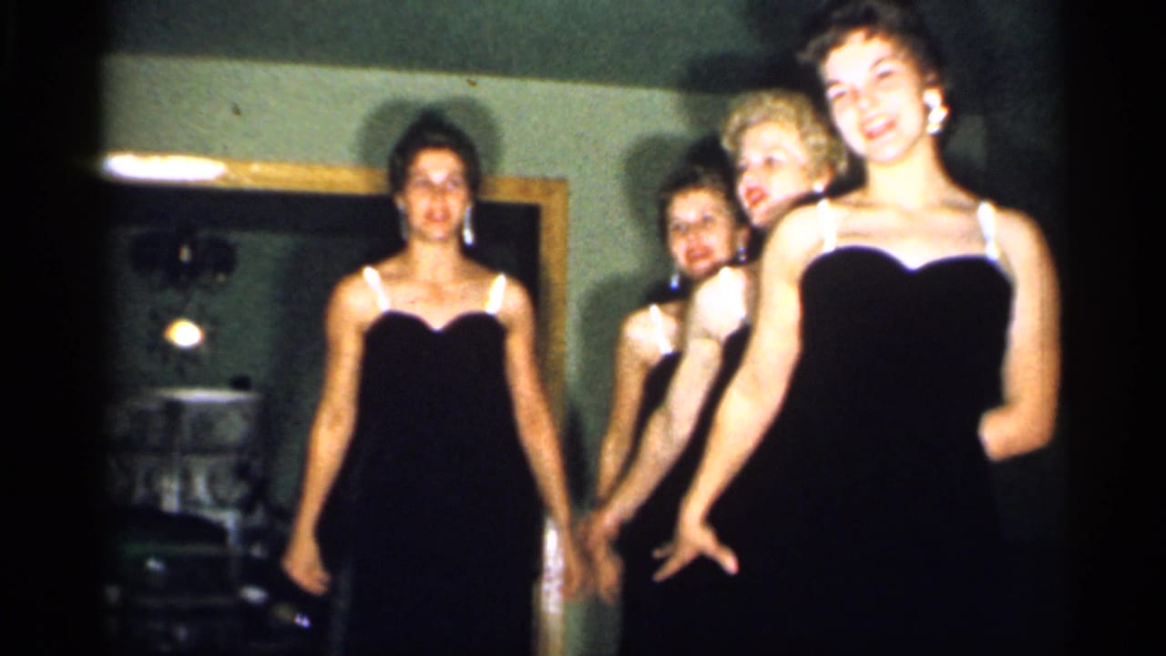 Four young women in matching dark dresses appear indoors in 1958 New Jersey home-movie footage, with a doorway or mirror behind them.