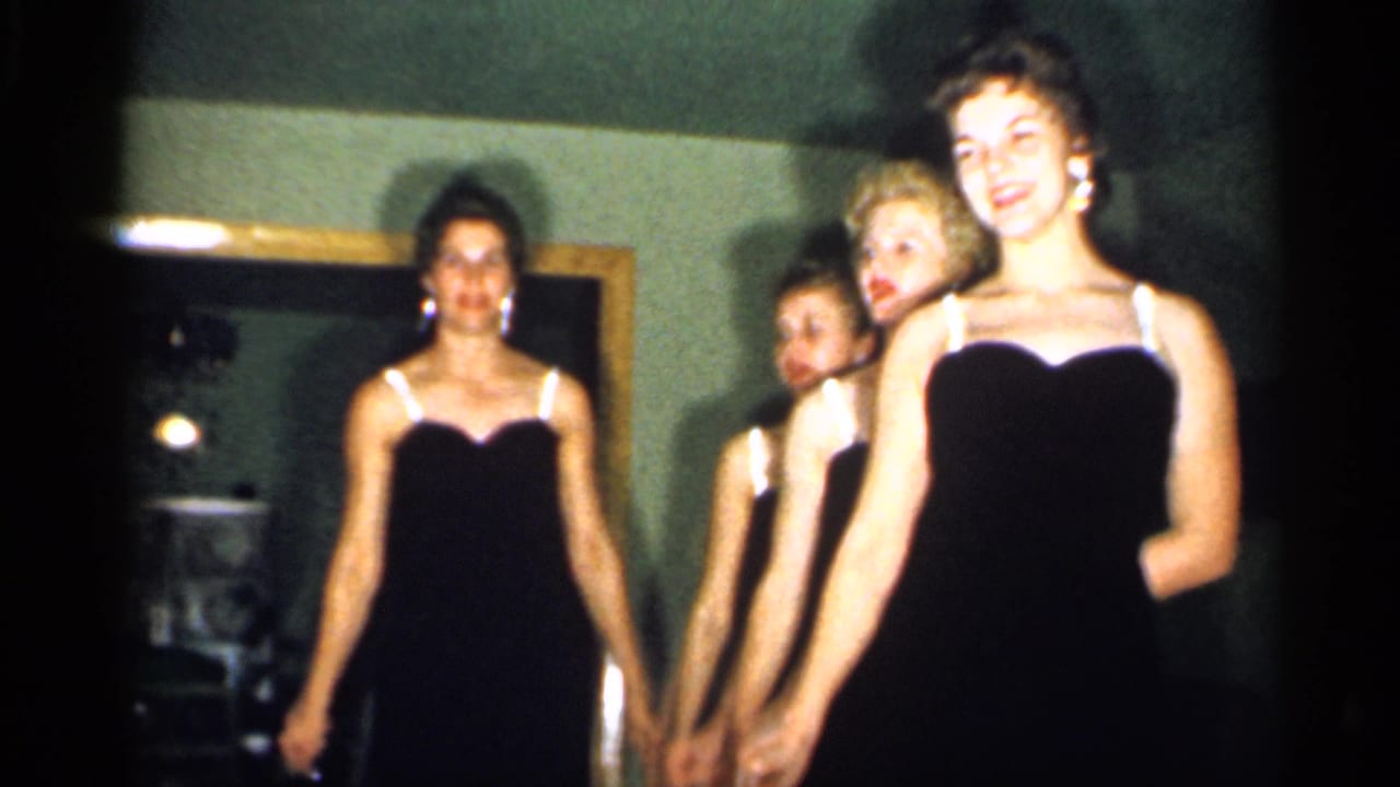 The women finish a synchronized movement in a dim private interior, with warm 8mm color and soft focus visible.