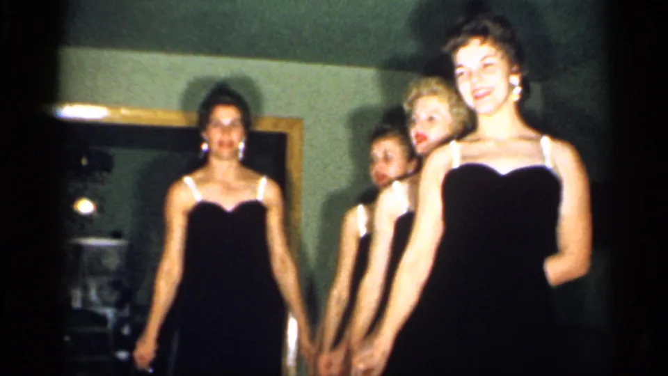The women finish a synchronized movement in a dim private interior, with warm 8mm color and soft focus visible.