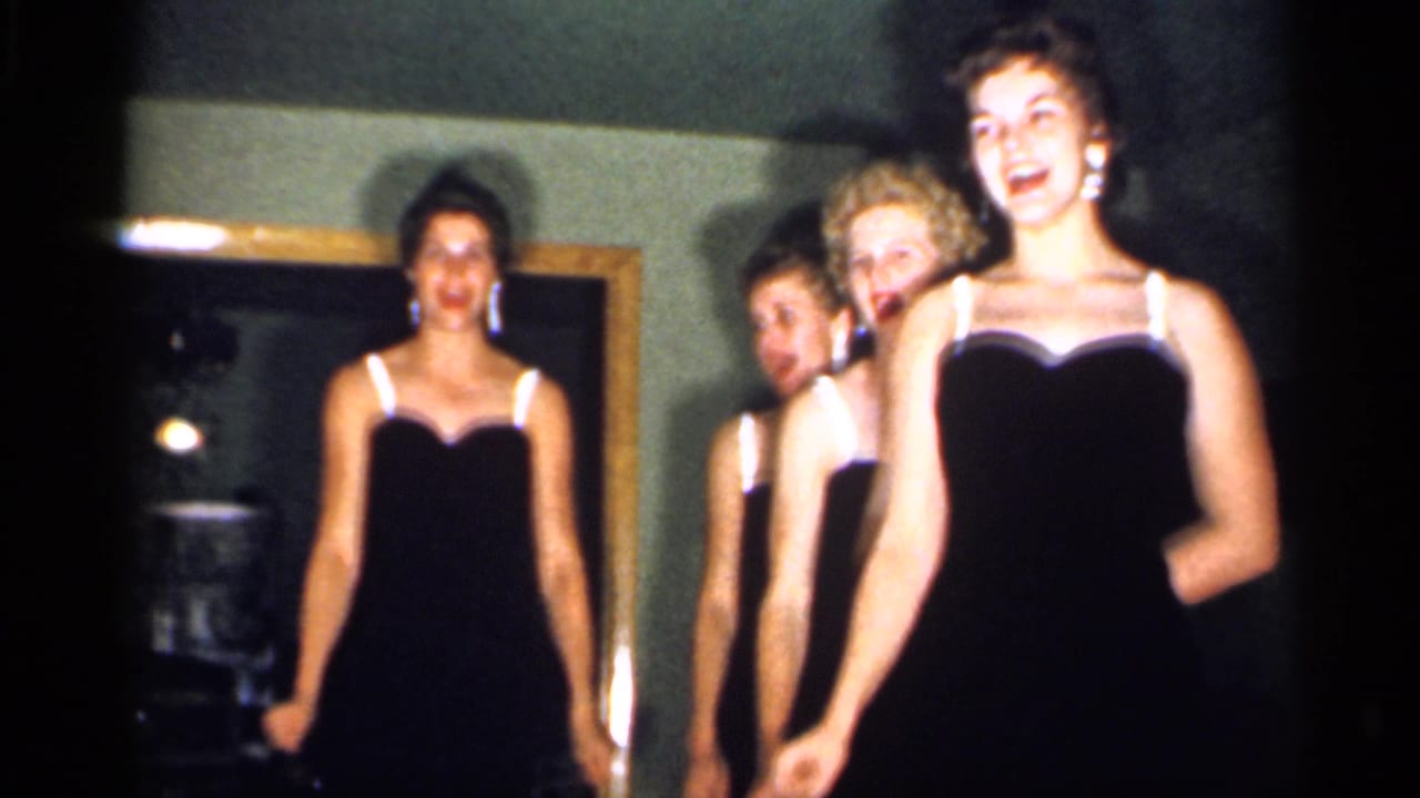 Four young women in black evening dresses with white straps appear to sing in a New Jersey living room in 1958 home-movie footage.