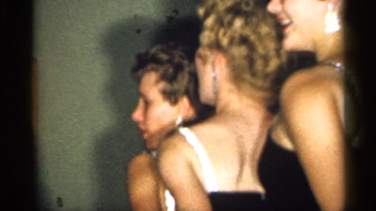 A close side view shows several women in dark evening dresses gathered indoors, with faces and backs partly visible in the home-movie frame.