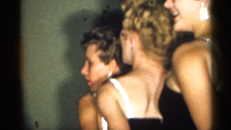 A close side view shows several women in dark evening dresses gathered indoors, with faces and backs partly visible in the home-movie frame.