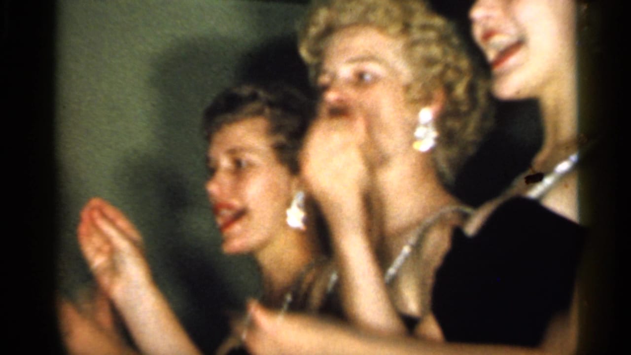 Young women in formal dresses laugh and clap at a 1958 New Jersey social event, with jewelry catching the light.