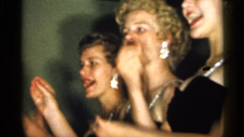 Young women in formal dresses laugh and clap at a 1958 New Jersey social event, with jewelry catching the light.