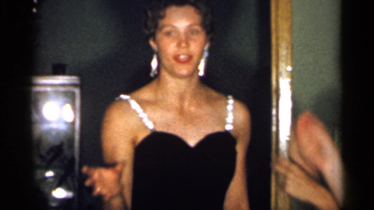 In 1958 New Jersey home-movie footage, a young woman in a black dress with bright straps gestures while facing the camera in a dim interior.