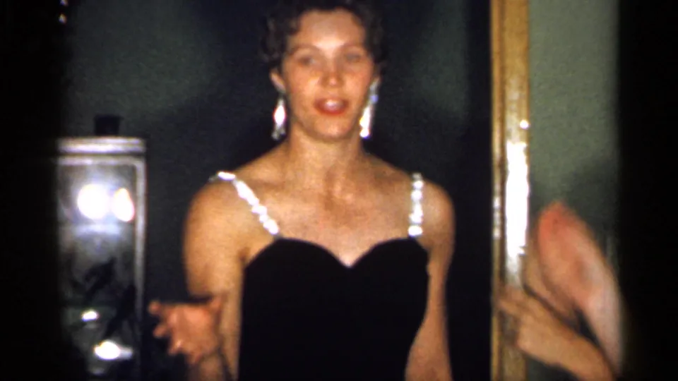 In 1958 New Jersey home-movie footage, a young woman in a black dress with bright straps gestures while facing the camera in a dim interior.