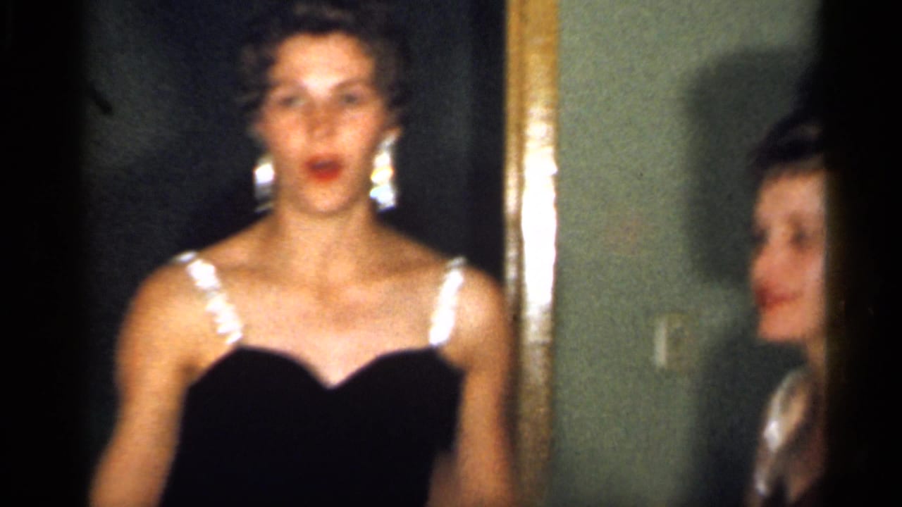 A young woman in a formal black dress stands by a framed doorway or mirror, with another woman's face partly visible at the right edge.