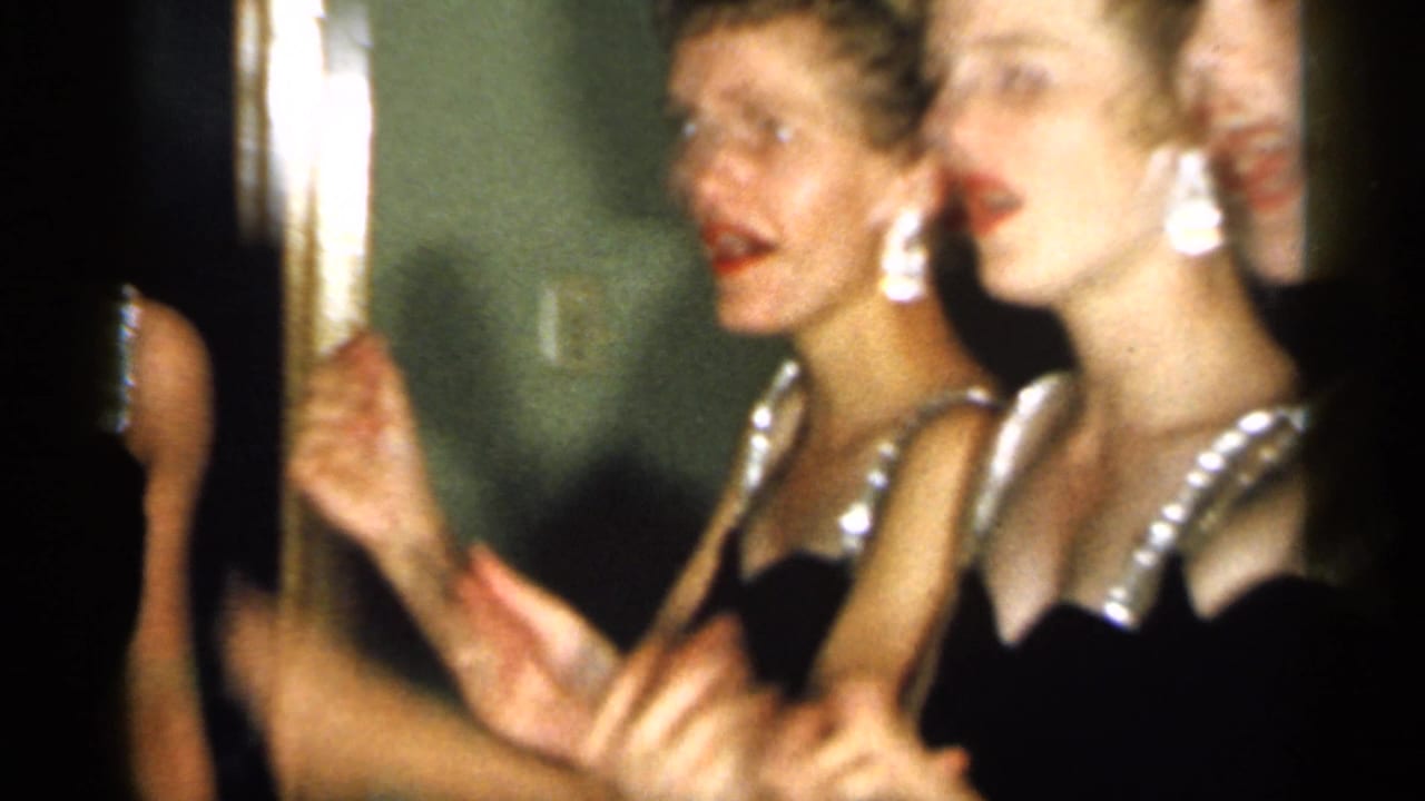 Two young women in black dresses with bright straps appear to sing or speak together, with motion-blurred hands in the foreground.