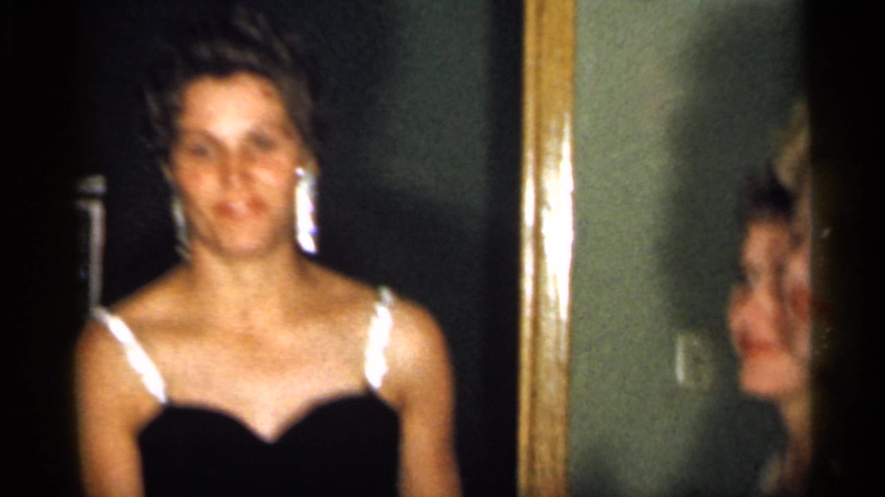 Young woman in a black dress with white straps stands before a mirror indoors.