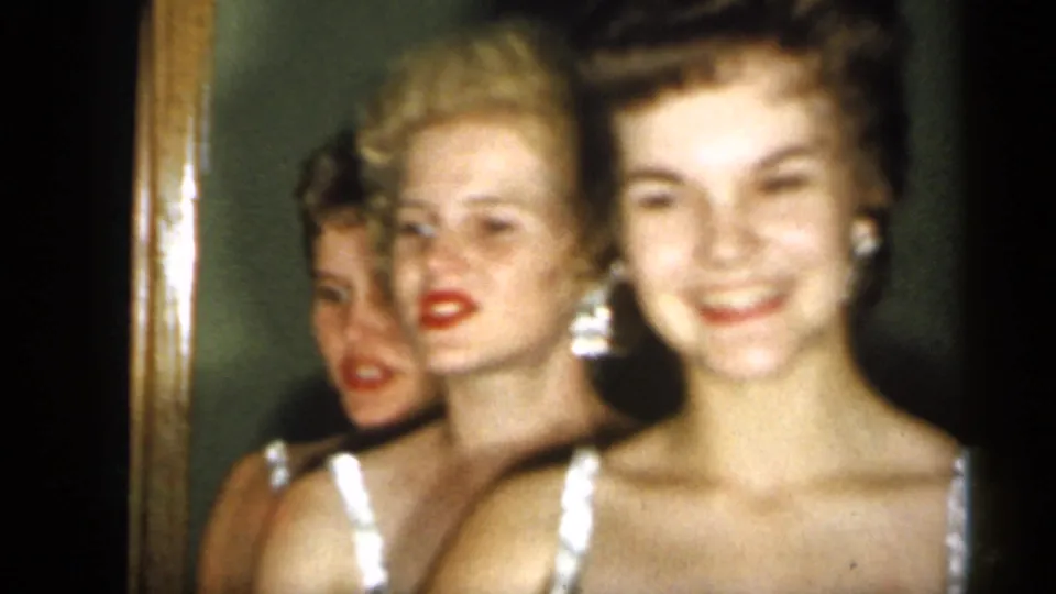 Several young women in black evening dresses and earrings smile close together beside a mirror in the 1958 New Jersey home-movie scene.