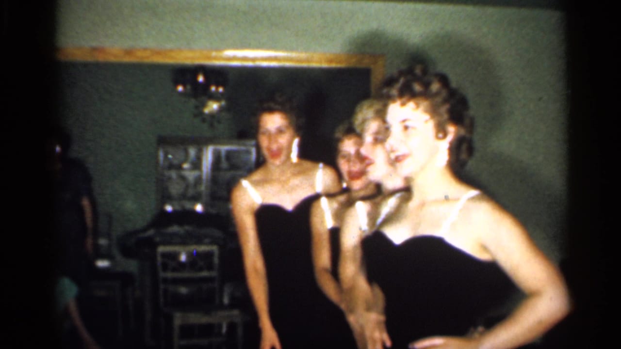 Young women in matching dark dresses turn and smile inside a home room.