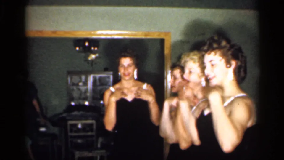 Several women gesture as if following directions or practicing a motion in the dim 1958 home-movie interior.