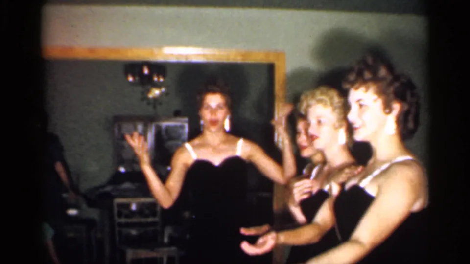 At the end of the clip, the women smile and gesture in coordinated dresses, with home furnishings and a chandelier behind them.
