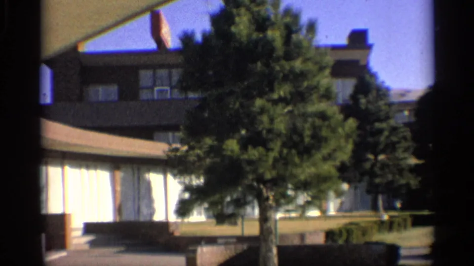 1959 Arizona home-movie frame showing a low-rise motel exterior with broad windows, lawn, trees, and blue sky.