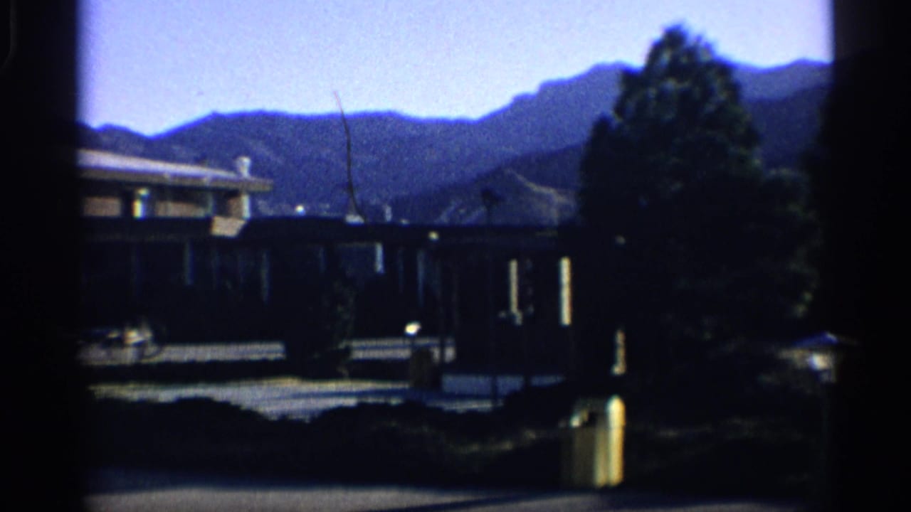 Low building and trees in front of a mountain range under blue sky.