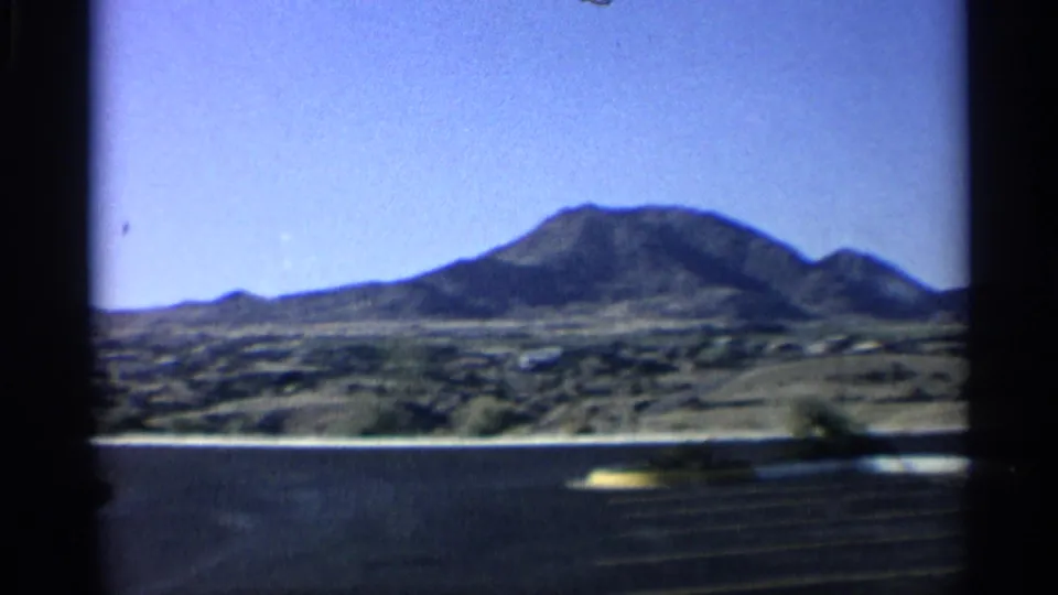 1959 Arizona 8mm landscape with a broad mountain, pale desert foreground, and distant low structures.
