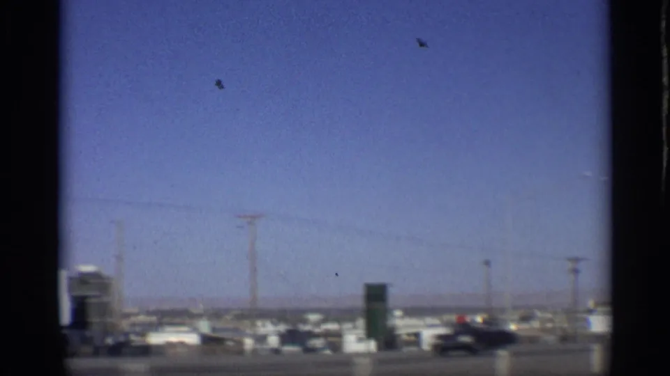 1959 Arizona highway view from a moving vehicle, with roadside signs, low buildings, utility poles and passing cars under a blue sky.