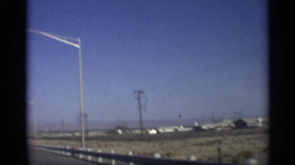 1959 Arizona home-movie frame from a moving vehicle showing roadside lights, utility poles, dry terrain, and low structures under blue sky.