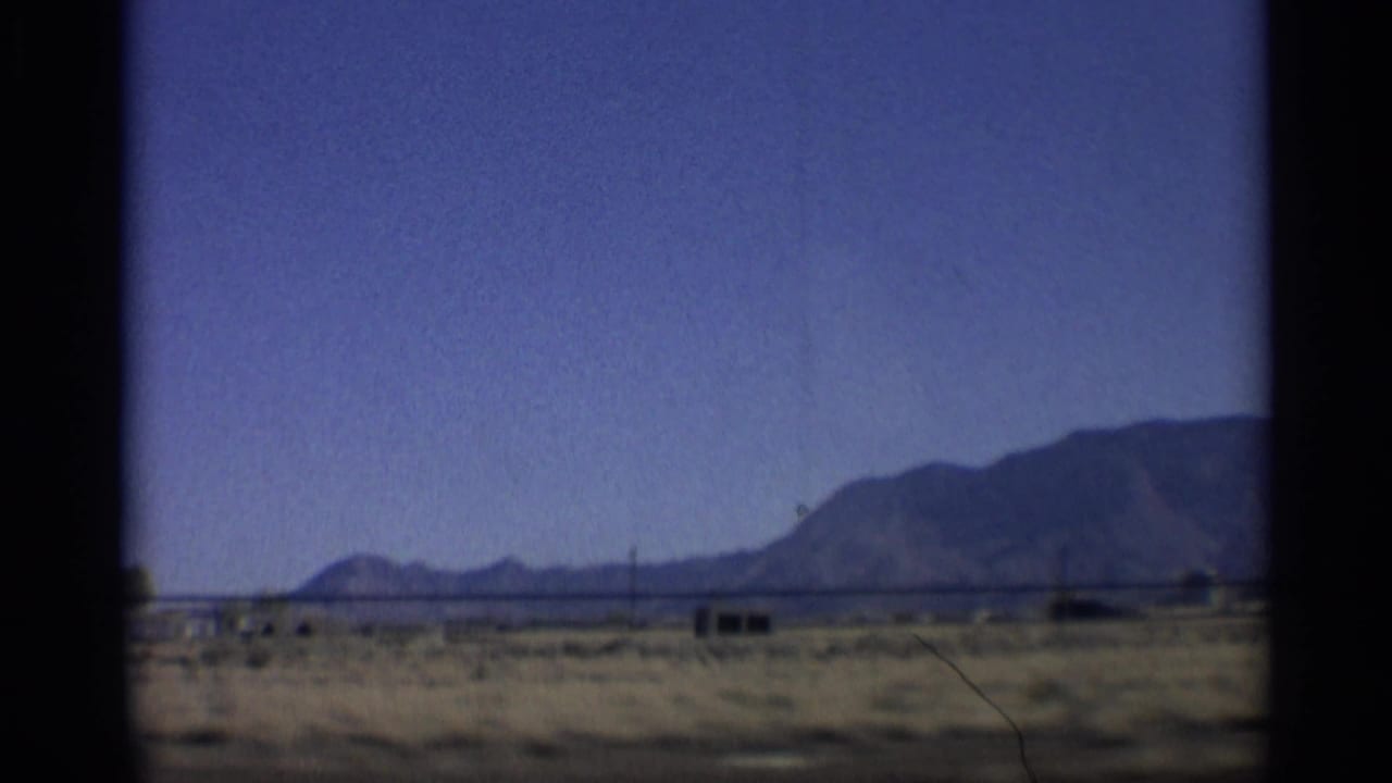 Open desert plain with distant mountains, small structures, utility poles, and blue sky.