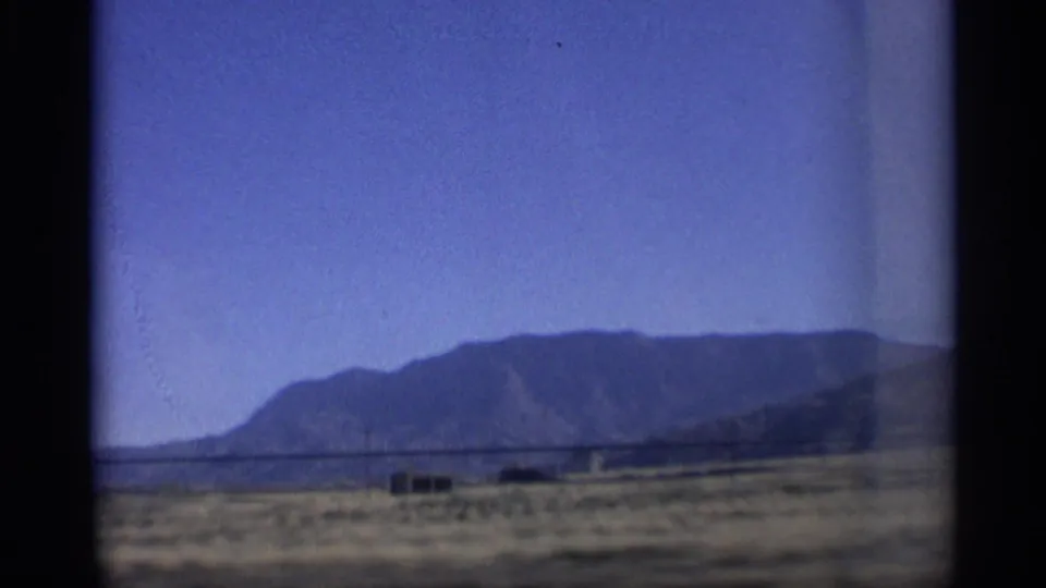 1959 Arizona home-movie frame showing a mountain ridge beyond dry roadside terrain, a small building, and utility poles.