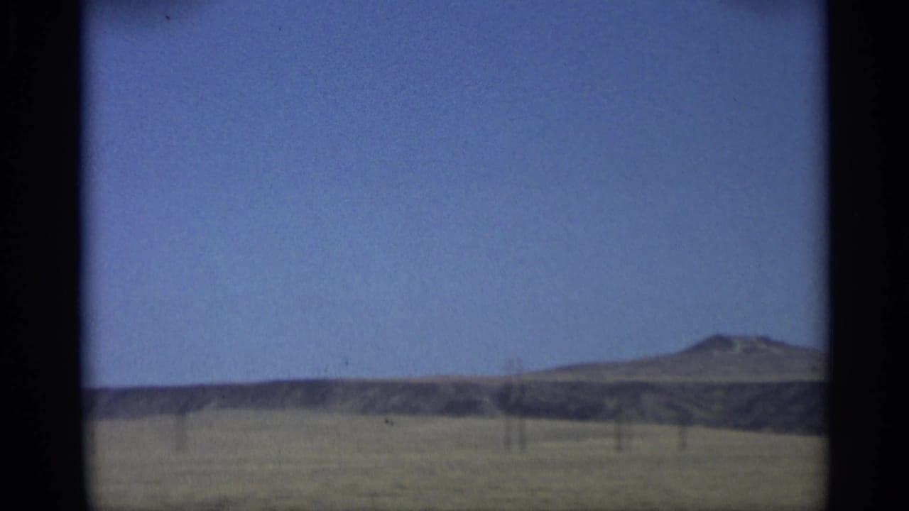 Distant small mountain and low ridge across open land beneath a blue sky.