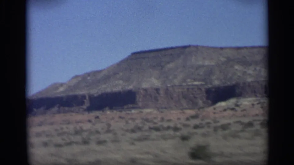 1959 Arizona home-movie frame showing a flat-topped mesa beyond scrubby desert foreground from a moving vehicle.