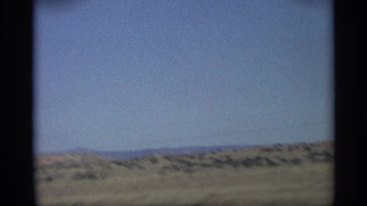 1959 Arizona home-movie frame showing open desert grassland, low ridges, and a broad blue sky from a moving viewpoint.