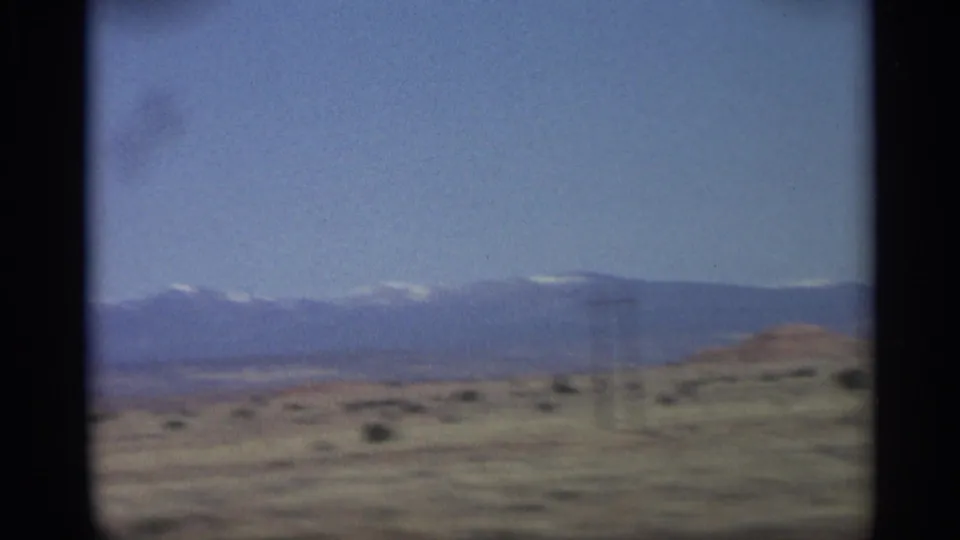 1959 Arizona road-view frame with arid foreground, distant mountains, pale snow-bright peaks, and a single utility pole.