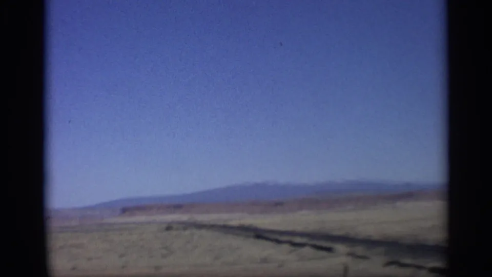 1959 Arizona home-movie view of flat desert terrain, a dark roadside strip, and low blue hills under clear sky.