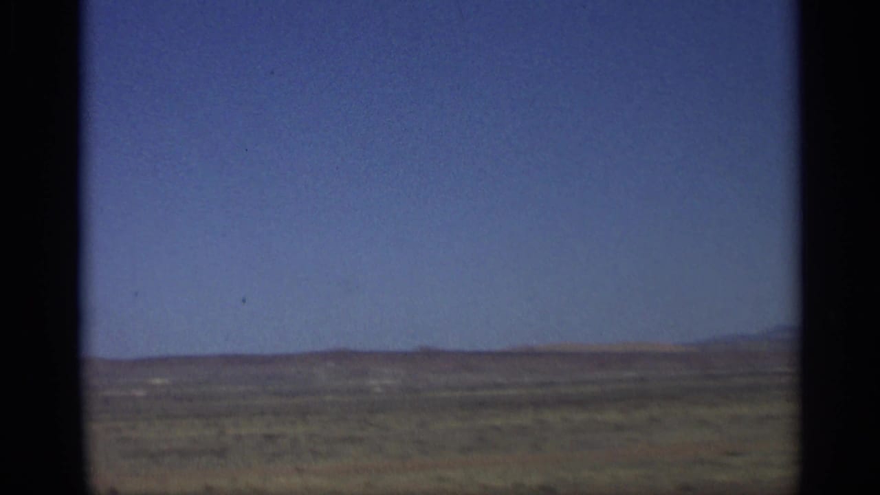 1959 Arizona home-movie frame showing open desert terrain, low distant hills, and a wide blue sky from a moving vehicle.