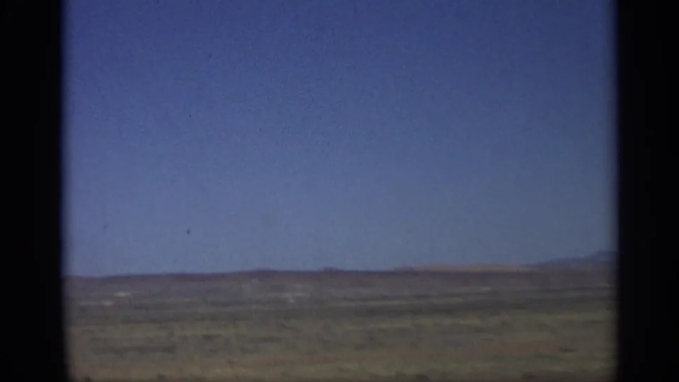 1959 Arizona home-movie frame showing open desert terrain, low distant hills, and a wide blue sky from a moving vehicle.